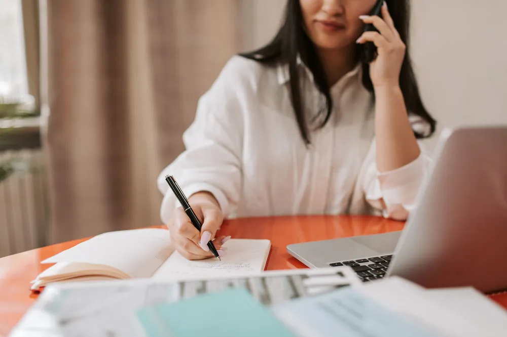 A woman on a phone call writing notes beside her laptop