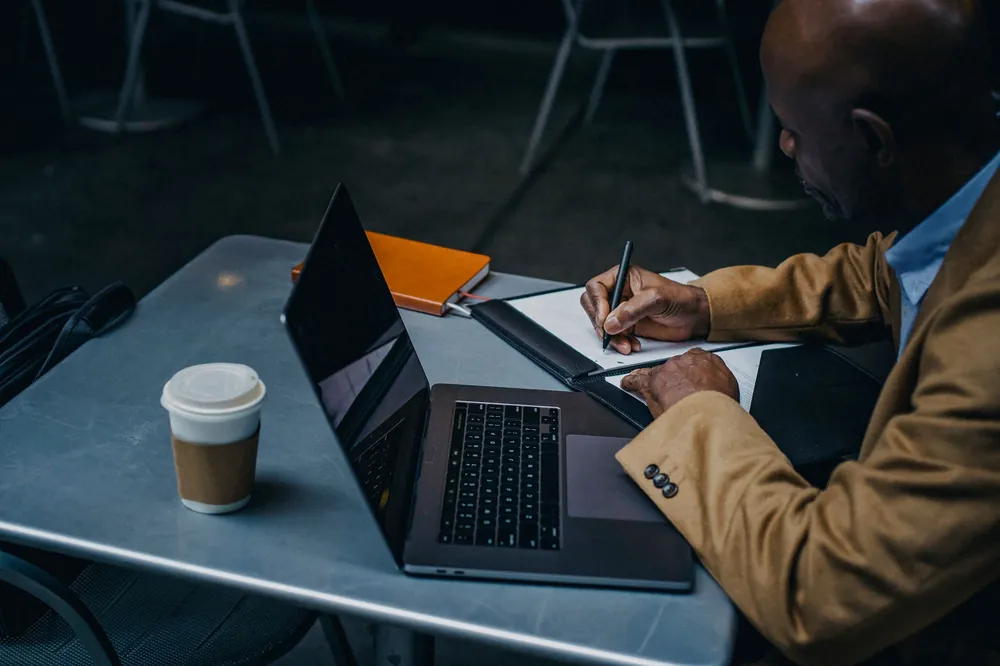 A person writing notes on paper beside a laptop and a coffee